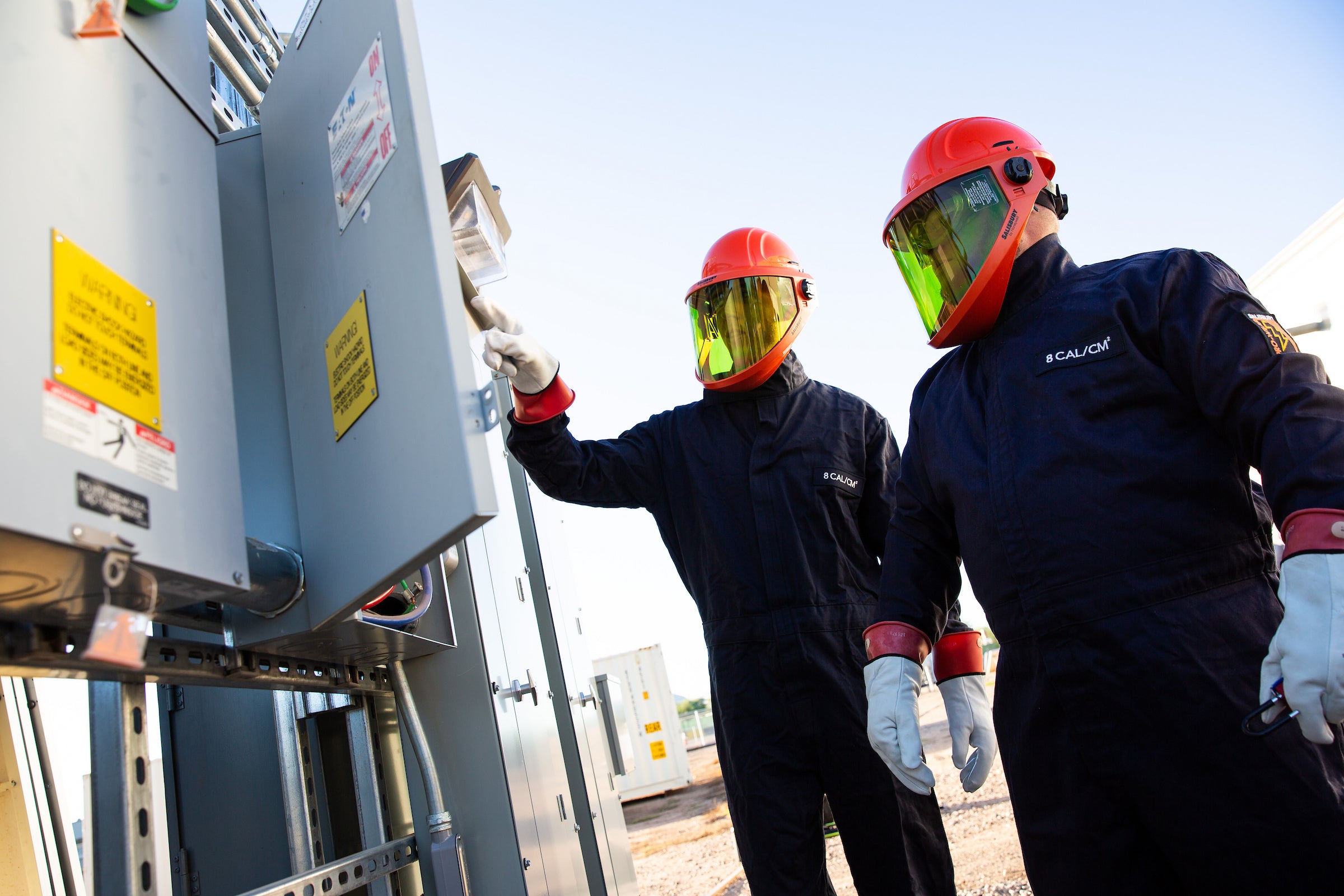 Two men wearing safety helmets and suits looking at an electric box.