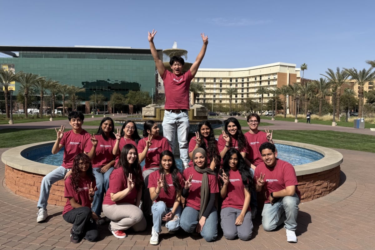 Group of people in maroon T-shirts posing in front of a fountain with buildings and palm trees in the background.