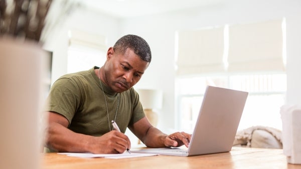 Man dressed in military T-shirt taking notes in front of a laptop.