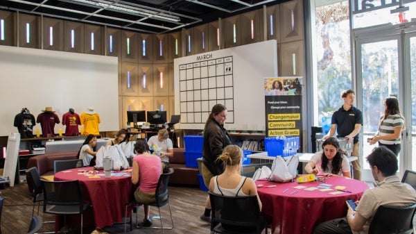 Students seated at round tables in a large room working together.