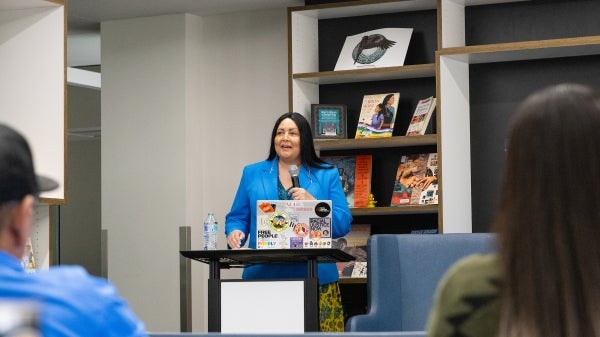 A  woman in a blue blazer speaks at a lectern with a laptop and bookshelves in the background.