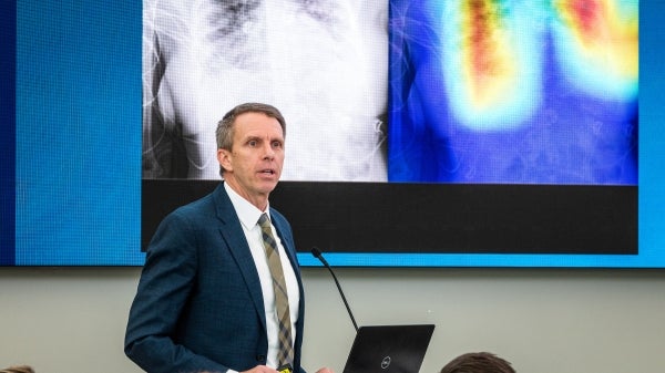 Man speaking into a microphone to an unseen audience in front of a screen displaying medical body scans.