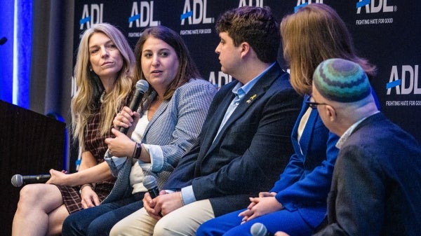 A panel of 5 people sit on stage while one holds a microphone and speaks