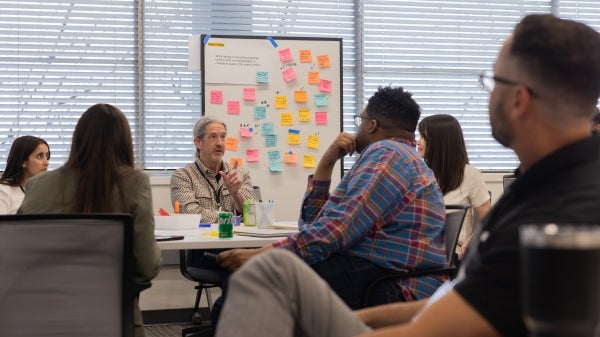 A seated man speaks to a room of people about his ideas with a whiteboard of sticky note ideas behind him. 