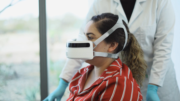 A woman wears the Breezing device on her face while a medical professional in a lab coat helps her.