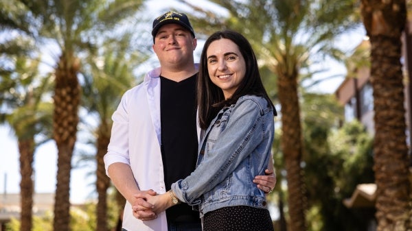 Man in black shirt and hat and woman with brown hair and jean jacket pose on Palm Walk holding hands