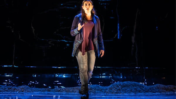 Woman with long dark hair dressed in casual clothes standing on a stage, gazing at the audience with one hand raised in a speaking gesture.