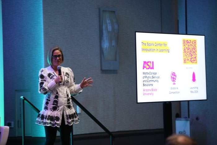 Woman stands on stage next to powerpoint on screen talking into microphone