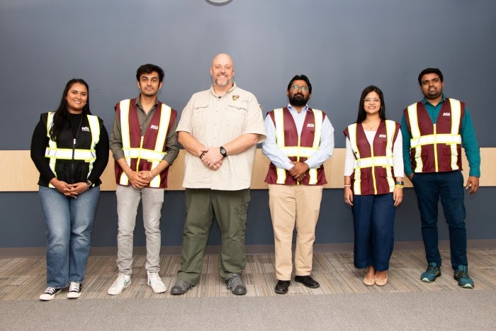 A group of six people stand in a line posing for a photo with some wearing safety vests