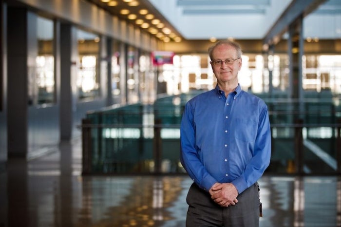 Bruce Rittmann, wearing a blue shirt and tan pants, stands in the hallway of the Biodesign Center with a glass window in the background. 