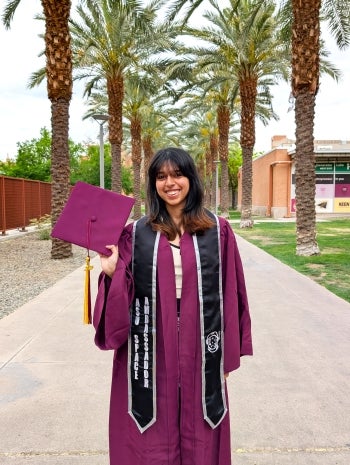 Ana Girish, 2025 spring grad, stands in her cap and gown on palm walk holding up her cap