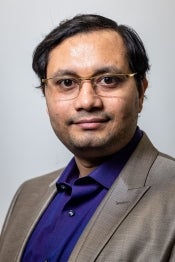 Portrait of a man with short black hair wearing glasses and a gray blazer with a blue button up shirt